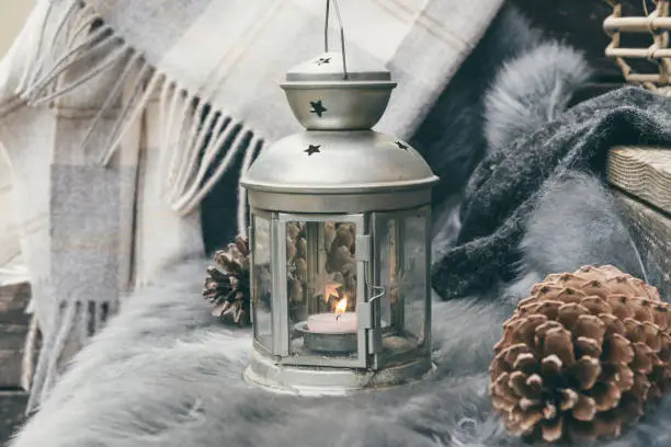 Cozy sitting area on wooden veranda with grey lambskin, lantern, fir cone and woolen scarf, grey colors