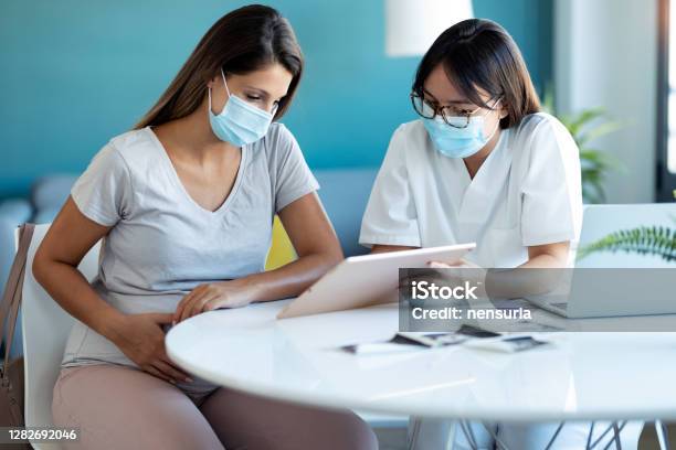 Young Beautiful Gynecologist Wearing A Hygienic Face Mask While Showing To Pregnant Woman Ultrasound Scan Baby With Digital Tablet In Medical Consultation Stock Photo - Download Image Now