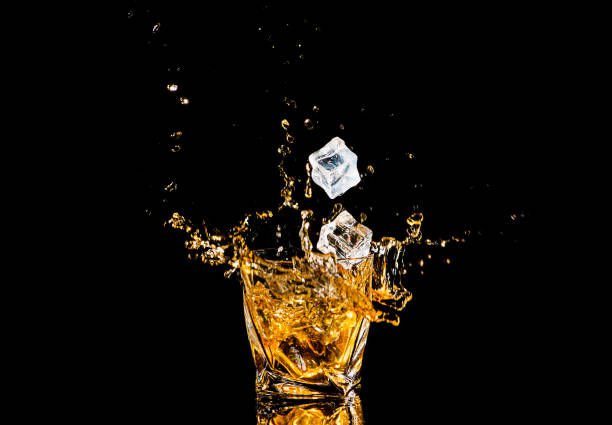 黒い背景の上に氷の立方体から飛び散ったウイスキーのグラス。アルコールが飛び散る。ウイスキーやコニャック、またはスプラッシュを持つアルコールの別のタイプ。 - whisky alcohol pouring glass ストックフォトと画像