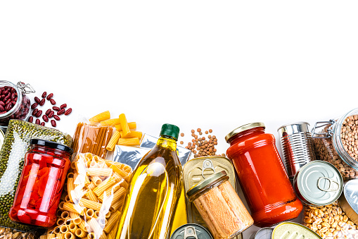 Top view of a group of multicolored non-perishable canned goods, conserves, sauces and oils arranged at the bottom of a white background making a frame and leaving useful copy space for text and/or logo. The composition includes cooking oil bottle, pasta, beans, preserves and tins. High resolution 42Mp studio digital capture taken with SONY A7rII and Zeiss Batis 40mm F2.0 CF lens