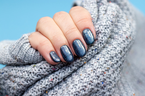 Female hand in a gray knitted sweater fabric with beautiful manicure - dark gray blue glittered nails. Selective focus. Closeup view