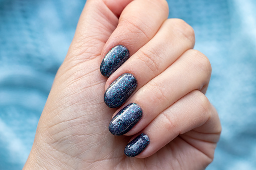 Female hand with beautiful manicure - dark gray blue glittered nails on blue knitted sweater fabric background. Selective focus. Closeup view