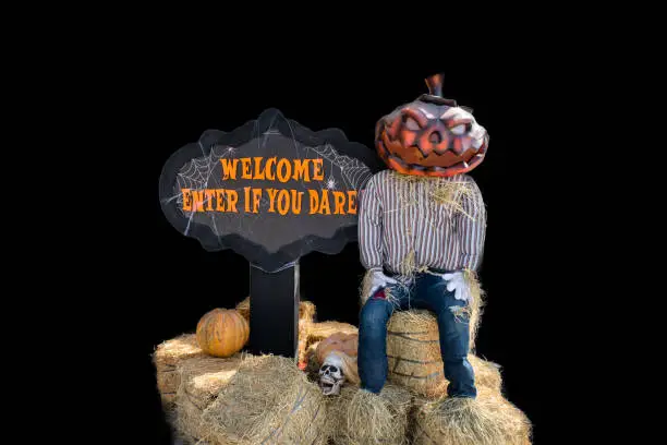 Photo of Halloween entrance decoration with spooky man with pumpkin head sitting on hay sheaves with sign 'Welcome, enter if you dare'