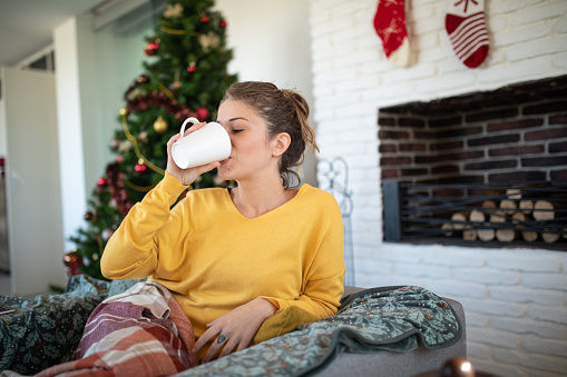 Young woman sitting on armchair and drinking hot drink on Christmas day at her cozy home