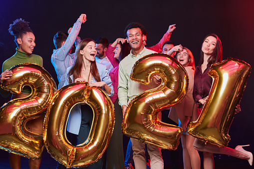 New Years Celebration. Group of young happy multiethnic people holding golden foil balloons in the form of numbers 2021 and having fun at Christmas party. Holiday concept