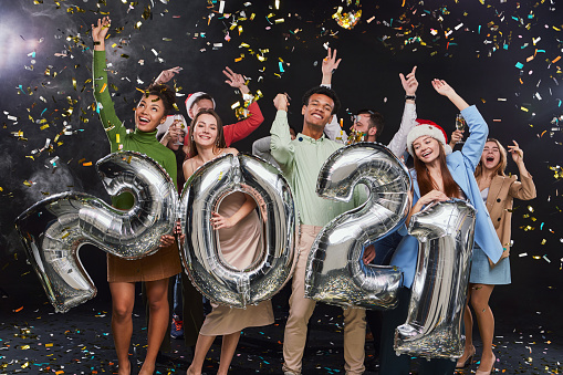 Group of young excited multiracial friends holding silver foil balloons in form of numbers 2021, raising hands up while celebrating New Years Eve, confetti falling in the air. Holiday, christmas party