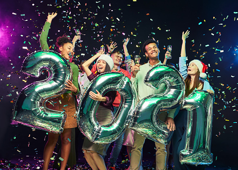 New Year celebration. Group of young happy multiracial friends holding silver foil balloons in form of numbers 2021, dancing and having fun, confetti falling in the air. People, party and holiday