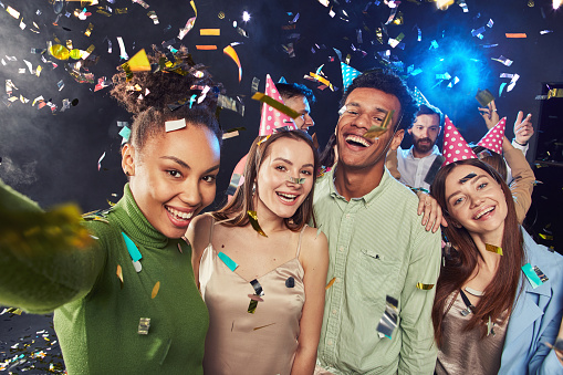 Group of young happy multiracial people wearing birthday hats making a selfie on mobile phone, confetti falling in the air. Holiday, party and celebration concept