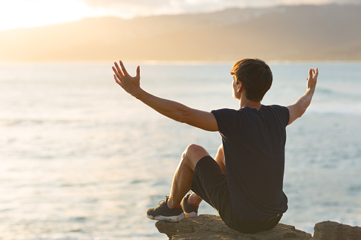 Free young male person feeling joyful. He is sitting alone on top of a mountain cliff facing a beautiful ocean view and raising both arms up to the sky.