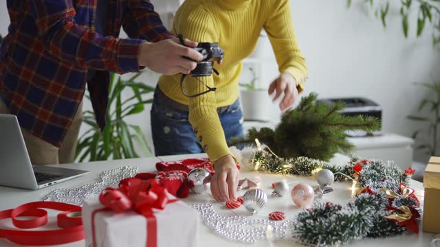 Tilt down of male photographer and female flatlay stylist discussing photos on camera while taking pictures of objects arranged on table to make Christmas still life images