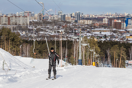Happy smiling man in a ski suit climbs the mountain on skis on a snow-covered slope using a drag lift. Over background of a coniferous forest and the cityscape.