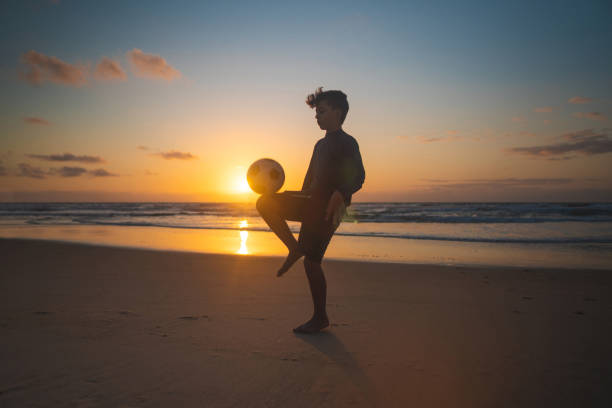 niño balanceando la pelota en la rodilla al amanecer - beach football fotografías e imágenes de stock