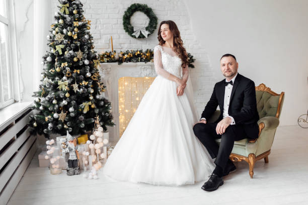 jovem casal apaixonado de noiva e noivo posando em estúdio ao fundo decorado com árvore de natal no dia do casamento no natal. aproveite um momento de felicidade e amor. - christmas wedding bride winter - fotografias e filmes do acervo