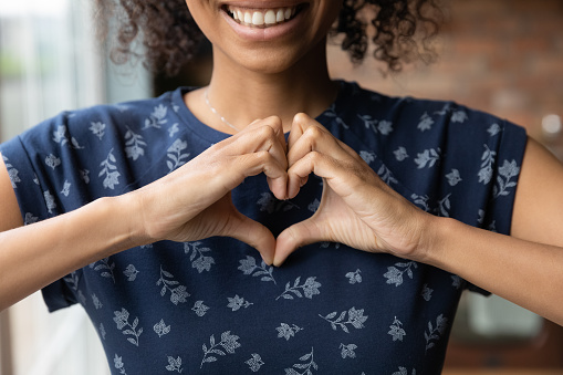 Crop close up of happy African American woman feel grateful thankful show heart sign spread love and care. Smiling biracial female volunteer make hand gesture support ill sick people patients.
