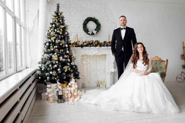 jovem casal apaixonado de noiva e noivo posando em estúdio ao fundo decorado com árvore de natal no dia do casamento no natal. aproveite um momento de felicidade e amor. - christmas wedding bride winter - fotografias e filmes do acervo