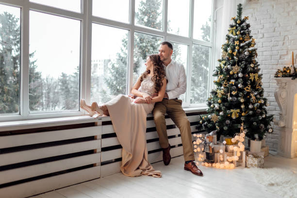 장식 된 스튜디오에서 큰 창문과 크리스마스 트리 근처에 앉아서 껴안고 축제 클러너드에 사랑에 커플. 행복한 휴가 - christmas wedding bride winter 뉴스 사진 이미지