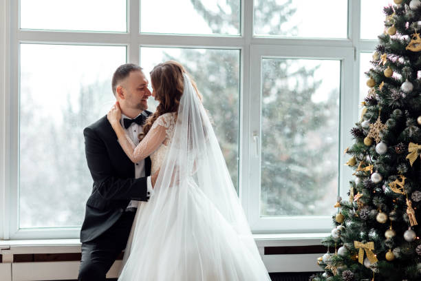 jovem casal apaixonado de noiva e noivo posando em estúdio ao fundo decorado com árvore de natal no dia de seu casamento no natal perto da grande janela panorâmica. - christmas wedding bride winter - fotografias e filmes do acervo