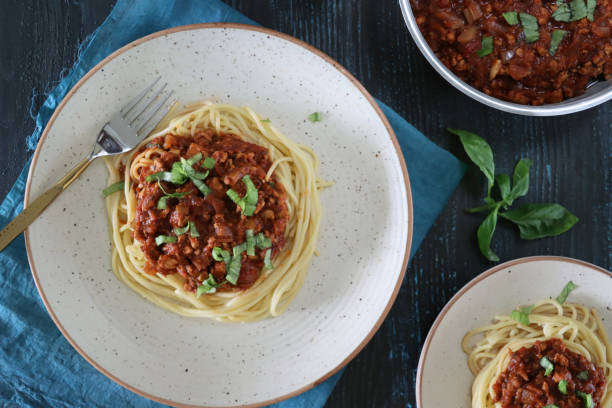 image of spaghetti pasta topped with homemade bolognese sauce of lean beef mince, rich tomato sauce, onions, garlic and basil leaf garnish, portion on white plate beside metal bowl containing bolognese sauce, elevate view - spaghetti cooked heap studio shot imagens e fotografias de stock
