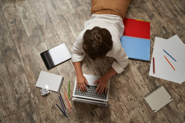 istock Little boy surfing internet or learning online, lying on the wooden floor at home and using laptop, top view. Happy childhood, elearning and distance education concept 1282727218