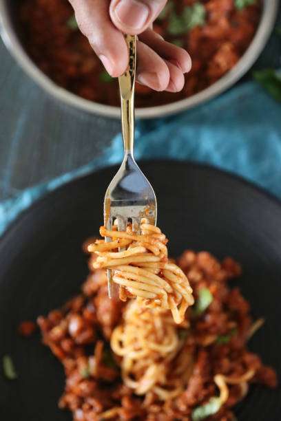 image of metal fork twirling spaghetti held in hand of unrecognisable person, pasta topped with homemade bolognese sauce of lean beef mince, rich tomato sauce, onions, garlic and basil leaf garnish, portion on black plate, elevated focus on foreground - spaghetti cooked heap studio shot imagens e fotografias de stock