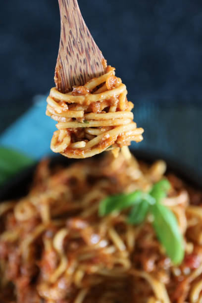 image of wooden fork twirling spaghetti pasta topped with homemade bolognese sauce of lean beef mince, rich tomato sauce, onions, garlic and basil leaf garnish, portion on black plate, elevated view, focus on foreground - spaghetti cooked heap studio shot imagens e fotografias de stock
