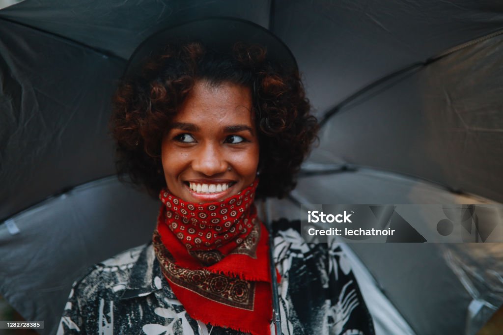 Portrait of a young black woman wearing a neck gaiter Young black woman walking on a rainy day, carrying an umbrella, wearing a bandana instead of the protective face mask in time of the coronavirus pandemic. Candid Stock Photo