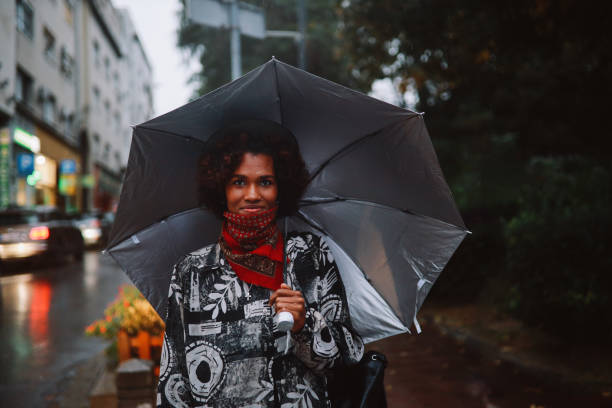 giovane donna di colore che cammina in una giornata piovosa indossando una ghetta al collo - candid women african descent umbrella foto e immagini stock