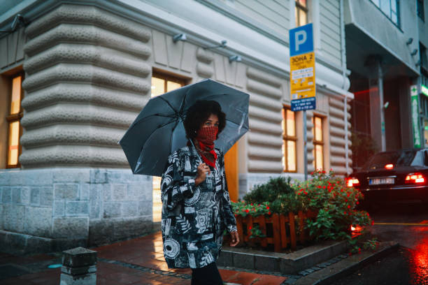 giovane donna di colore che cammina in una giornata piovosa indossando una ghetta al collo - candid women african descent umbrella foto e immagini stock