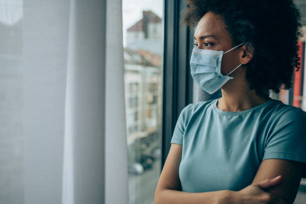 Worried young woman looking through window at home in quarantine. Beautiful african-american woman wearing protective face mask and looking through window at home during Coronavirus/COVID-19 pandemic. epidemic stock pictures, royalty-free photos & images