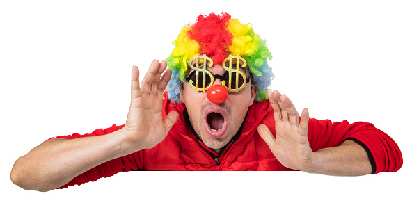 Funny clown in a multi colored wig leaning over a white blank sign holding his hands near mouth while telling something. He is wearing golden dollar shaped sunglasses. Isolated on white. You can add extra white space with your message to the bottom.