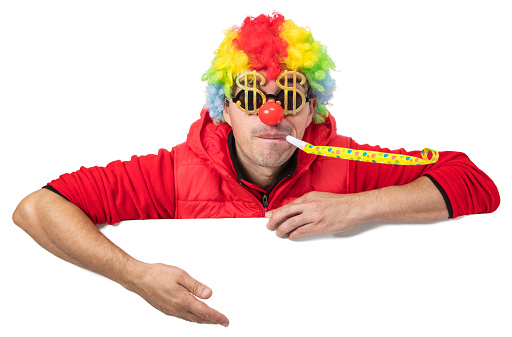 Funny clown in a multi colored wig with dollar shaped glasses and horn blower holding a blank white sign showing something. Isolated on white. You can add extra white space with your message to the bottom.