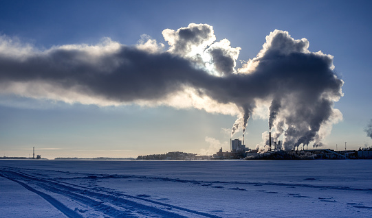 View of factory power plant in winter with steam rising into deep blue sky, sun shines behind steam, sea port with the boat at horizon line