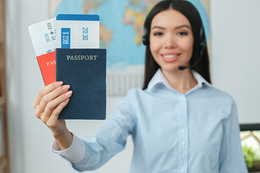 Young woman travel agent in tour agency standing wearing headset holding passports and tickets close-up