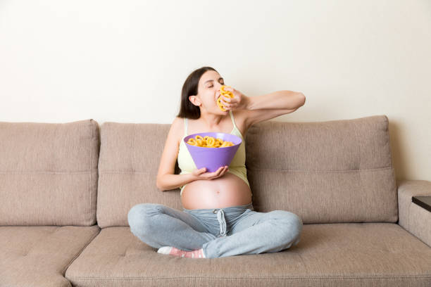 Pregnant woman sitting on the sofa enjoys eating chips from a bowl at home. Feeling hungry during pregnancy concept Pregnant woman sitting on the sofa enjoys eating chips from a bowl at home. Feeling hungry during pregnancy concept. family eating potato chips stock pictures, royalty-free photos & images