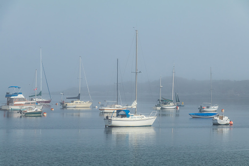sailboat in dense fog bank with smaller row boat towed behind obscured and barely visible in dense fog bank on the bay water of Bahia de San Quintin, on the Pacific coast of Baja, Mexico