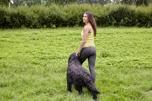Back view on the black briard and his young female owner who are walking on the grass in the public park.