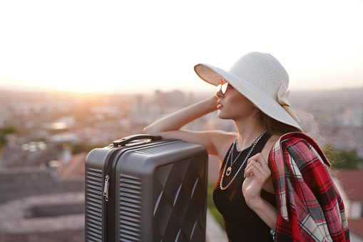 smiling blonde woman in hat traveling in tourist city with suitcase.