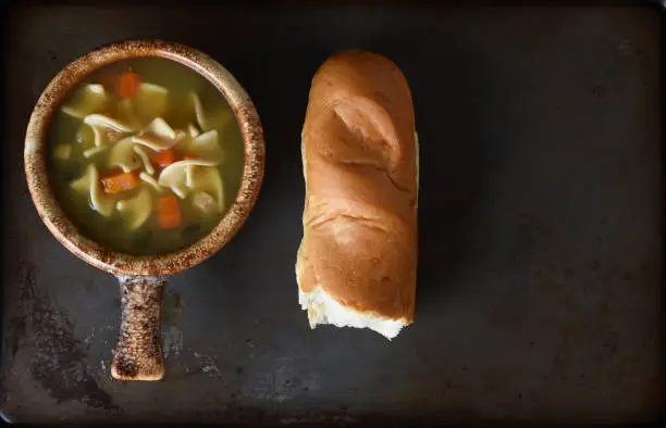 A bowl of Chicken Noodle Soup and a baguette on a metal baking sheet on a rustic wood kitchen table.