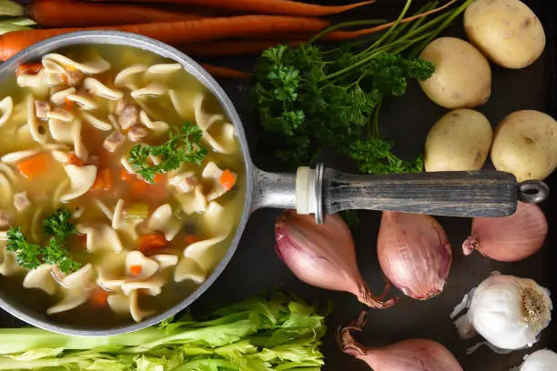 A pot of fresh homemade Chicken Noodle Soup surrounded by some of the ingredients.