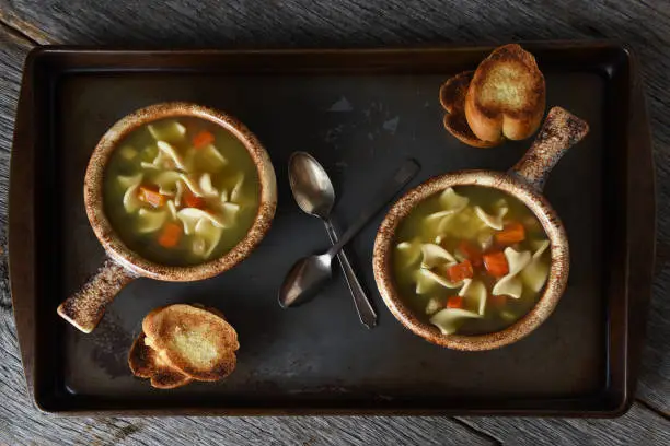 Two bowls of Chicken Noodle Soup, a Baguette  Garlic Toast and spoons on a metal baking sheet on a rustic wood kitchen table.