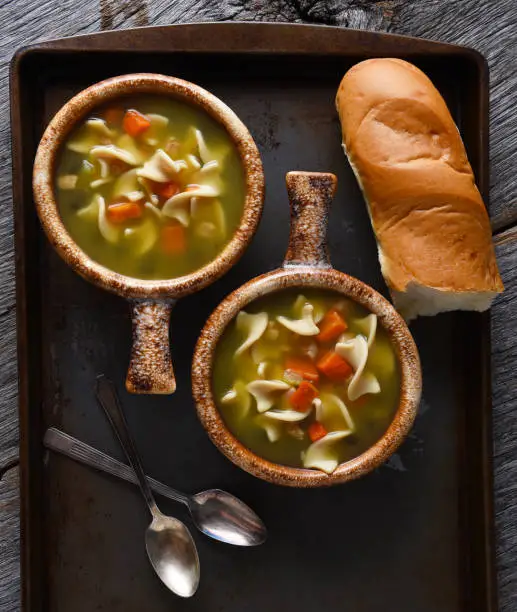 Two fresh homemade bowls of Chicken Noodle Soup with a baguette and spoons. Closeup in vertical format.
