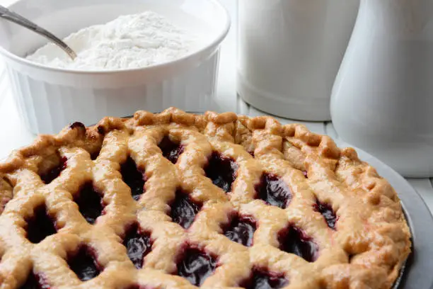 High angle shot of a fresh baked cherry pie with a lattice crust. A bowl of flour and pitchers fill the background. Horizontal format.