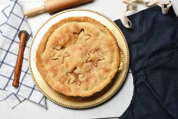 Fresh baked homemade apple pie on a white wood table with apron, rolling pin and crimper.