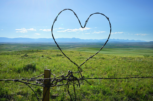 A heart made of barbed wire on the top of a fence in Montana, in front of a grassland scene with mountains in the distance on a sunny day.
