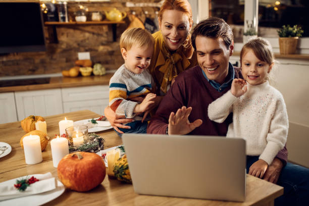 Happy family having video call before Thanksgiving meal at home. Happy parents and kids using computer and waving while making video call berofe Thanksgiving dinner in dining room. virtual event photos stock pictures, royalty-free photos & images