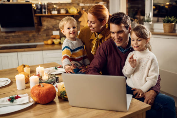 Happy parents and kids using laptop before Thanksgiving dinner in dining room. Happy family making video call over laptop while celebrating Thanksgiving at home. thanksgiving holiday covid stock pictures, royalty-free photos & images