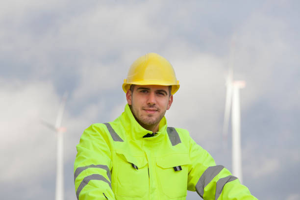 istock Smiling young engineer with hard hat and protective clothing in front of blurred wind turbines in the background 1282793184