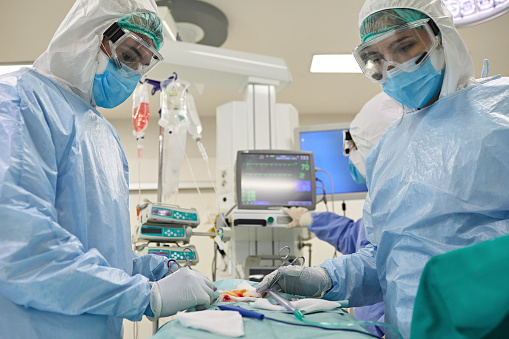 Doctors wearing protective suits, operating gowns, surgical masks, eyewear, and gloves working in operating room.