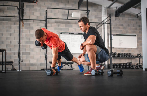 Early 30s Male Athlete Doing Renegade Row Push-Ups at Gym Fitness instructor watching strong sportsman use dumbbells to work his back, biceps, core, and shoulders during push-ups. fitness instructor stock pictures, royalty-free photos & images