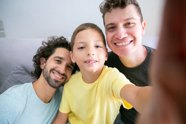 Selfie of cheerful homosexual dads and kid sitting on couch Selfie of cheerful homosexual dads and kid sitting on couch at home and smiling at frontal camera. Front view. Family and communication concept gay daddy stock pictures, royalty-free photos & images